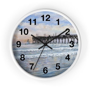 May include: A white round clock with black numbers and hands. The clock face features a painting of a wooden pier extending over a blue ocean with white waves. A fishing boat is visible in the distance.