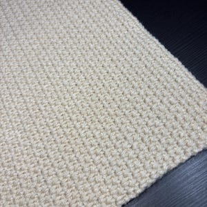 May include: Close-up of a cream-colored, hand-crocheted fabric. The texture is dense and uniform, with visible stitches creating a subtle pattern. The yarn appears soft and the overall look is cozy and handmade.