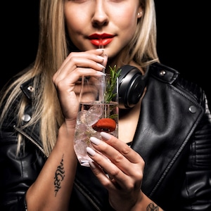 Puede incluir: Una mujer con una chaqueta de cuero negra sostiene un vaso de ginebra y tónica con hielo y una ramita de romero. La bebida está adornada con una fruta roja.