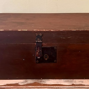 May include: A rectangular wooden chest with a dark brown finish. The chest has a metal latch and lock on the front. The wood shows signs of wear, suggesting it is an antique or vintage piece. The chest is sitting on a light pink table.