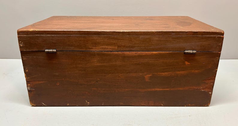 May include: A rectangular wooden box with a hinged lid. The box is stained a dark brown color, with visible wood grain and some wear. The hinges are metal. The box is likely used for storage.
