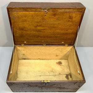 May include: An open, antique wooden chest with a hinged lid. The chest is brown with a lighter interior and brass hardware. The lid is held open by two cords. The chest is empty.
