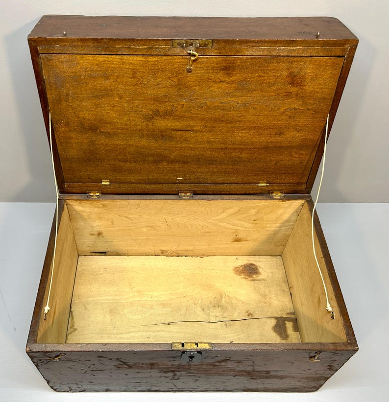 May include: An open, antique wooden chest with a hinged lid. The chest is brown with a lighter interior and brass hardware. The lid is held open by two cords. The chest is empty.
