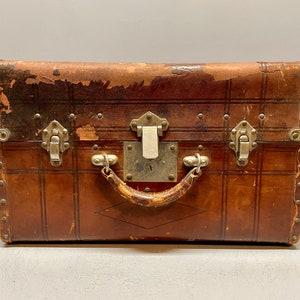 May include: A vintage brown leather suitcase with metal hardware, including clasps and a handle. The suitcase has a worn and distressed look, adding to its antique charm.