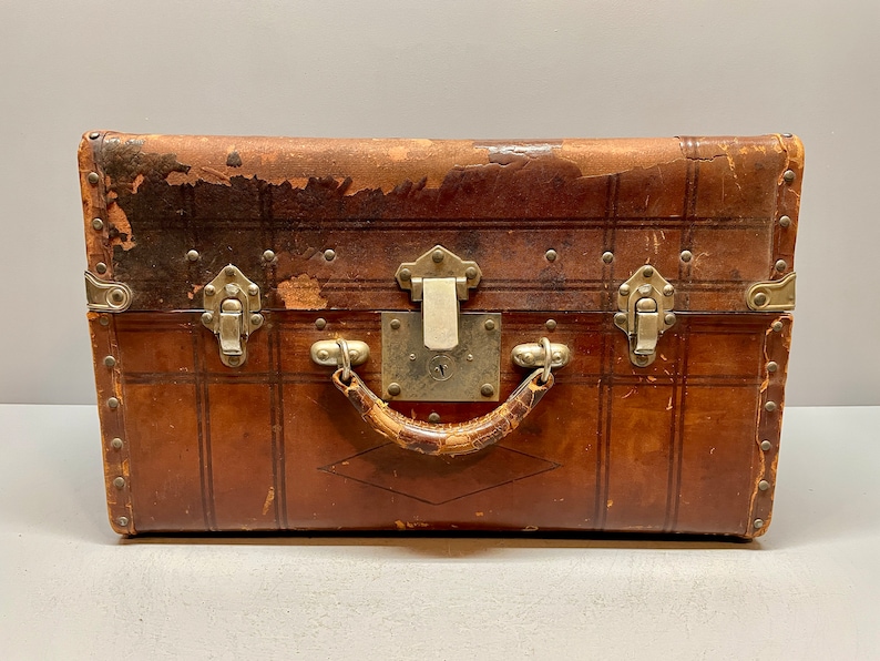 May include: A vintage brown leather suitcase with metal hardware, including clasps and a handle. The suitcase has a worn and distressed look, adding to its antique charm.