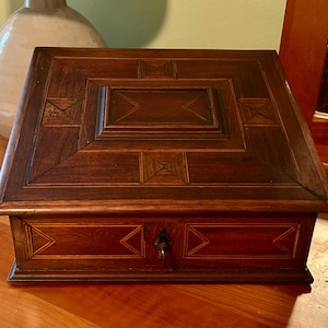May include: A dark brown wooden box with a geometric inlaid design on the lid and front. The box has a rectangular shape with a hinged lid and a small keyhole on the front. The box is sitting on a wooden surface.