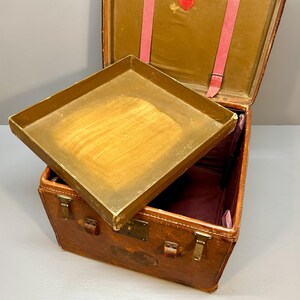 May include: An antique brown leather trunk with a hinged lid and a removable tray. The trunk has metal clasps and corner protectors. The interior is lined with a burgundy fabric. Pink straps are visible inside the open lid.