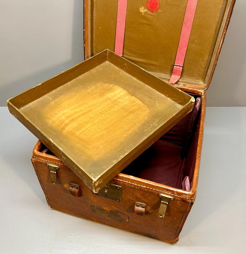 May include: An antique brown leather trunk with a hinged lid and a removable tray. The trunk has metal clasps and corner protectors. The interior is lined with a burgundy fabric. Pink straps are visible inside the open lid.