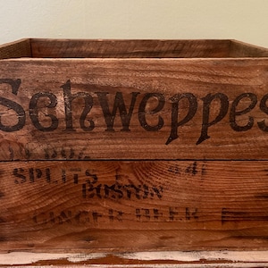 May include: Vintage wooden crate with the word "Schweppes" printed in black. The crate is made of weathered wood and has additional text that is difficult to read. The crate is a square shape and is likely used for storage or display.