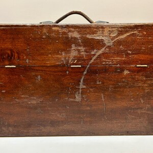 May include: A vintage wooden toolbox with a dark brown finish. The box has a leather handle and metal clasps. The wood shows signs of wear, including scratches and imperfections, adding to its antique charm.