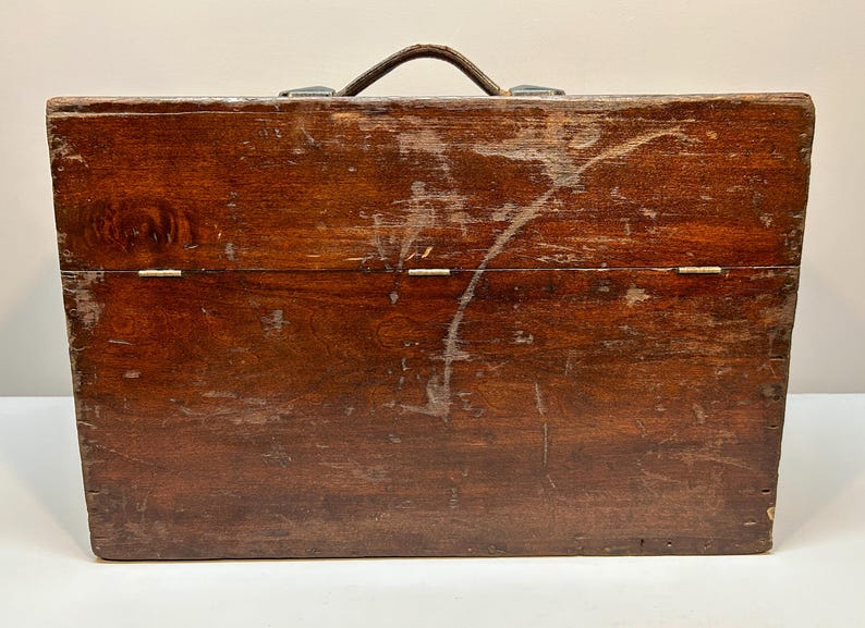 May include: A vintage wooden toolbox with a dark brown finish. The box has a leather handle and metal clasps. The wood shows signs of wear, including scratches and imperfections, adding to its antique charm.