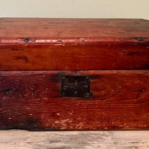 May include: A weathered, rectangular wooden chest with a hinged, curved lid. The chest is stained a deep reddish-brown color, with visible wood grain and signs of wear. A metal latch secures the front.