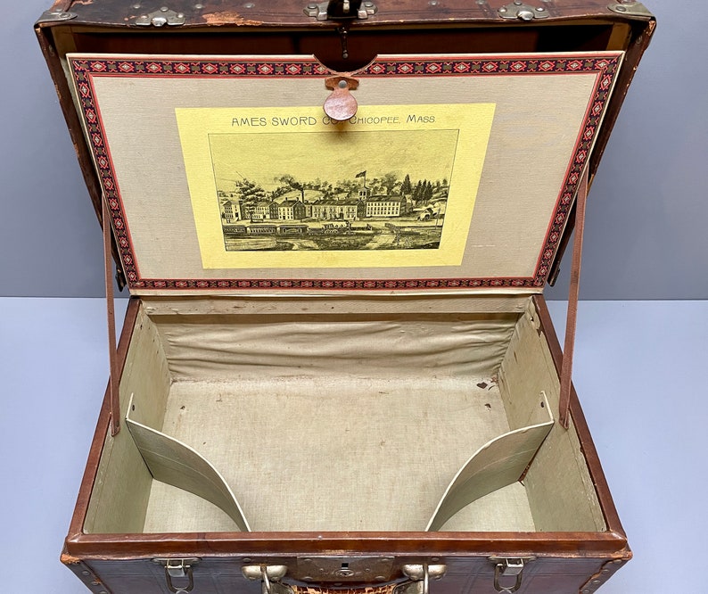 May include: An antique brown leather suitcase with brass hardware and a fabric lining. The inside of the suitcase has two compartments. The suitcase has a label that reads "AMES SWORD CO. CHICOPEE MASS."