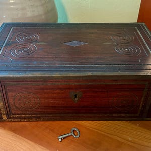 May include: A rectangular wooden box with a dark finish, decorative brass accents, and a keyhole. The lid features carved spiral and diamond designs. A small key rests in front of the box. The box is likely used for storage.