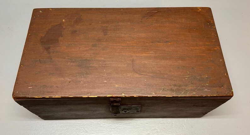 May include: A rectangular wooden box with a hinged lid and a metal clasp. The box is stained a dark brown color, showing the wood grain. The box appears to be vintage or antique, with some wear and tear visible on the surface.