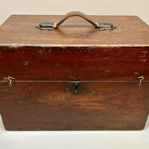 May include: A rectangular wooden box with a leather handle and metal hardware. The box is stained a dark brown color and has a keyhole on the front. The box is likely used for storage.