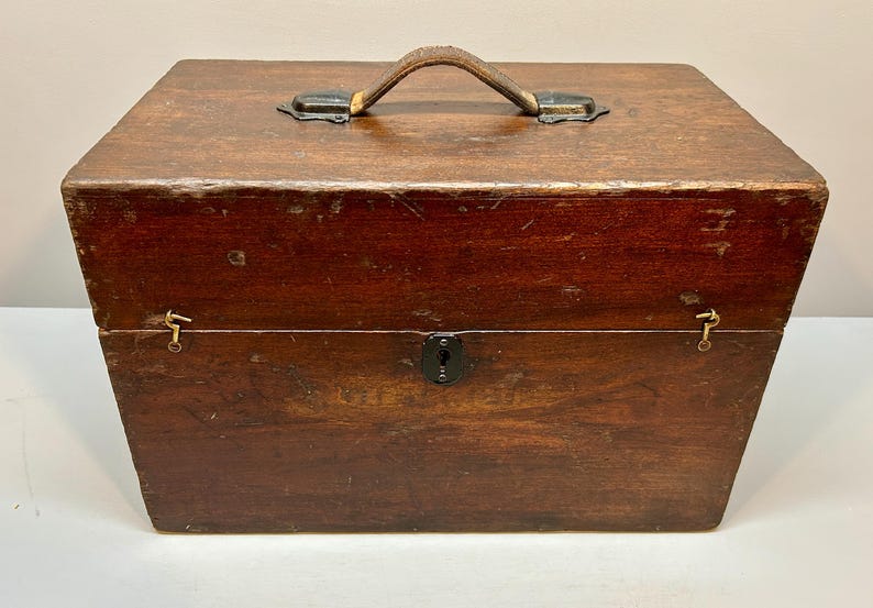 May include: A rectangular wooden box with a leather handle and metal hardware. The box is stained a dark brown color and has a keyhole on the front. The box is likely used for storage.