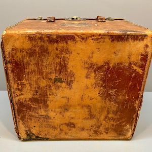 May include: A vintage, rectangular leather trunk with a worn, distressed appearance. The trunk is a warm, reddish-brown color with visible scratches and wear. It features a metal clasp and handle on top, and decorative stitching along the edges.