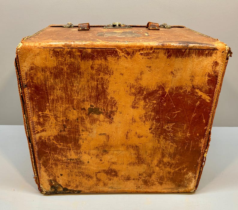 May include: A vintage, rectangular leather trunk with a worn, distressed appearance. The trunk is a warm, reddish-brown color with visible scratches and wear. It features a metal clasp and handle on top, and decorative stitching along the edges.