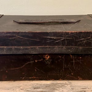 May include: A rectangular, dark brown antique box with a leather handle on the top. The box has metal trim and a keyhole on the front. The box appears to be made of wood and leather, and shows signs of wear.