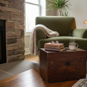 May include: Cozy living room scene with a green armchair, a wooden trunk used as a side table, and a fireplace. A stack of books, eyeglasses, and a steaming mug sit on the trunk. A blanket drapes over the chair.