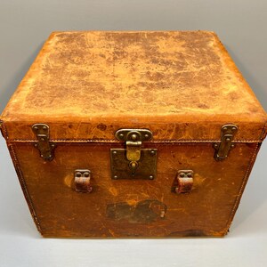 May include: A vintage, rectangular trunk with a weathered, light brown leather exterior. The trunk features brass hardware, including a central lock and corner protectors. The aged leather shows signs of wear, adding to its antique charm.