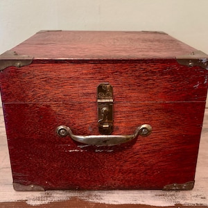 May include: A rectangular wooden box with a deep reddish-brown finish, featuring brass-toned metal corner protectors, a central brass latch mechanism, and a curved metal handle attached to the front panel with visible screws.