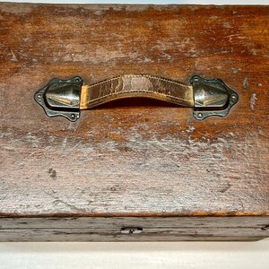May include: A vintage wooden box with a distressed finish. The box features a leather handle with metal hardware. The wood has a rich, warm brown color with visible grain and wear, suggesting age and use. The box is rectangular with a hinged lid.