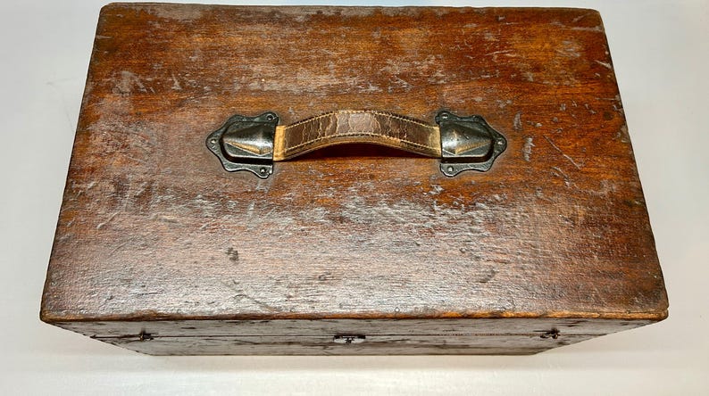 May include: A vintage wooden box with a distressed finish. The box features a leather handle with metal hardware. The wood has a rich, warm brown color with visible grain and wear, suggesting age and use. The box is rectangular with a hinged lid.