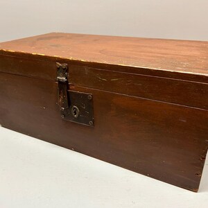 May include: A rectangular wooden chest with a hinged lid and a metal clasp. The chest is stained a dark brown color, with visible wood grain. The metal clasp and keyhole are aged, adding to the vintage aesthetic. The chest is likely used for storage.
