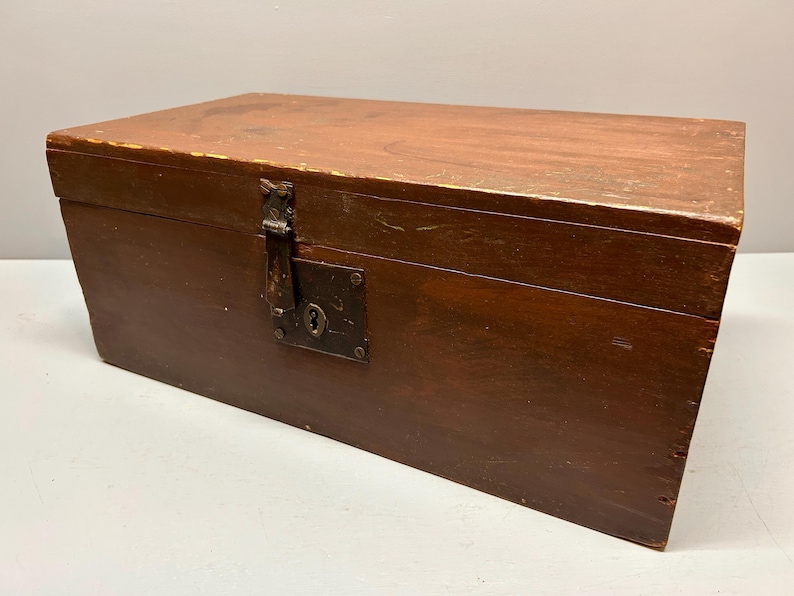 May include: A rectangular wooden chest with a hinged lid and a metal clasp. The chest is stained a dark brown color, with visible wood grain. The metal clasp and keyhole are aged, adding to the vintage aesthetic. The chest is likely used for storage.