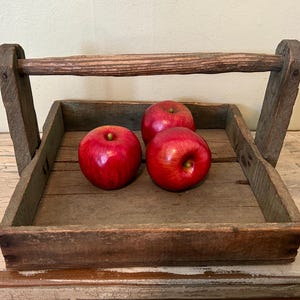May include: A rustic wooden tray with a handle, perfect for carrying or displaying items. The tray is filled with four red apples.