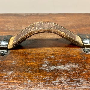 May include: Close-up of a vintage wooden trunk with a worn, brown leather handle. The handle is attached to the trunk with black metal hardware. The wood has a rich, aged appearance.