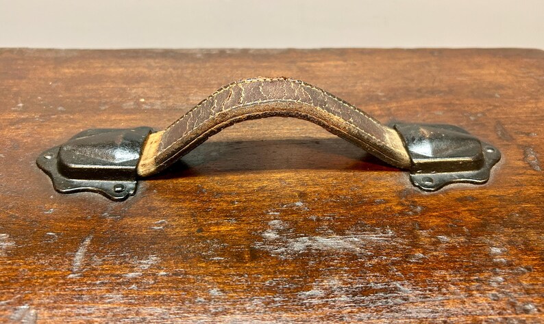 May include: Close-up of a vintage wooden trunk with a worn, brown leather handle. The handle is attached to the trunk with black metal hardware. The wood has a rich, aged appearance.