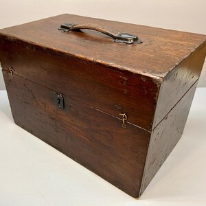 May include: A rectangular wooden box with a leather handle and metal hardware. The box is stained a dark brown color and has a keyhole on the front. The box appears to be vintage or antique.