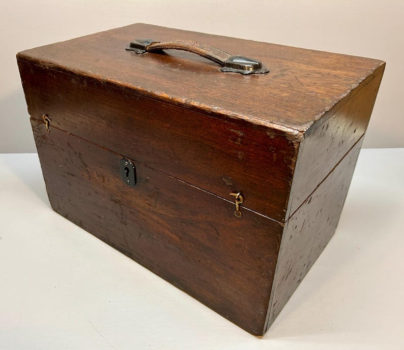May include: A rectangular wooden box with a leather handle and metal hardware. The box is stained a dark brown color and has a keyhole on the front. The box appears to be vintage or antique.
