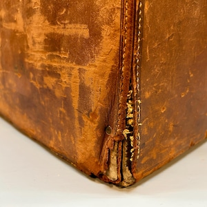 May include: Close-up of a vintage brown leather trunk. The trunk shows signs of wear, with visible stitching and aged leather. The corners are reinforced, and the overall appearance suggests a well-traveled piece.