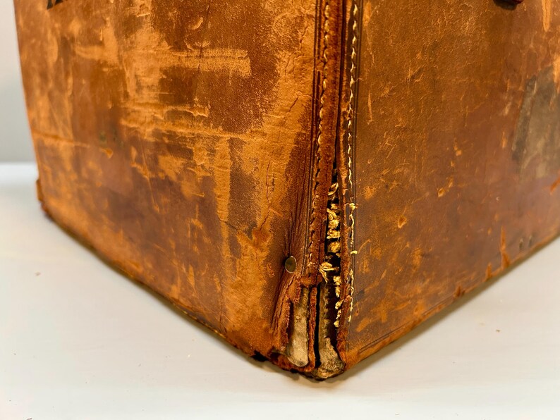 May include: Close-up of a vintage brown leather trunk. The trunk shows signs of wear, with visible stitching and aged leather. The corners are reinforced, and the overall appearance suggests a well-traveled piece.