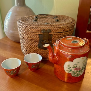 May include: An orange ceramic teapot and two matching cups with floral designs. The teapot has a lid and spout. A woven basket and a large vase are in the background. The tea set is on a wooden surface.