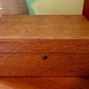 May include: A rectangular wooden box with a hinged lid. The box is made of a reddish-brown wood with visible grain. The box is sitting on a wooden surface. The box has a small keyhole on the front.