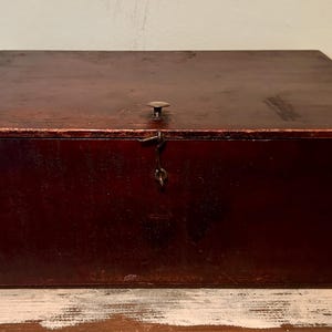 May include: A rectangular, antique wooden box with a dark reddish-brown finish. The box has a hinged lid with a small metal knob and a latch. The box is likely used for storage and sits on a distressed wooden surface.