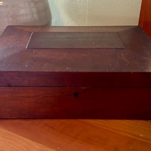 May include: A rectangular, dark brown wooden box with a hinged lid. The box has a keyhole and is sitting on a wooden surface. The lid has a recessed panel in the center. The box is likely used for storage.