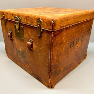 May include: A vintage, square-shaped trunk made of distressed brown leather. The trunk features brass hardware, including a lock and corner protectors. The initials "H.D.K." are stenciled on the side. The trunk has leather handles and is likely used for storage.