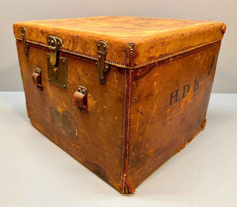 May include: A vintage, square-shaped trunk made of distressed brown leather. The trunk features brass hardware, including a lock and corner protectors. The initials "H.D.K." are stenciled on the side. The trunk has leather handles and is likely used for storage.