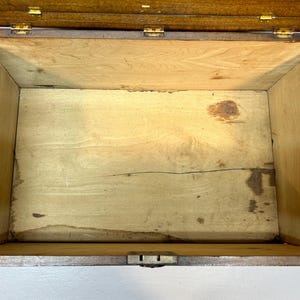 May include: An open, antique wooden box with a hinged lid. The interior is light-colored wood, showing signs of age and wear. The box has brass hinges and a latch, and is suspended by two cords.