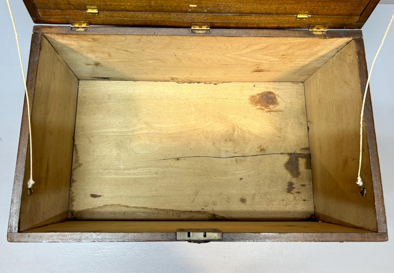 May include: An open, antique wooden box with a hinged lid. The interior is light-colored wood, showing signs of age and wear. The box has brass hinges and a latch, and is suspended by two cords.