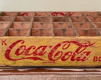 May include: Vintage wooden Coca-Cola bottle crate with a yellow and red painted design. The crate has multiple compartments and metal corner reinforcements. The words "DRINK Coca-Cola IN BOTTLES" are printed on the front.