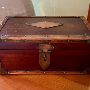 May include: A rectangular wooden chest with a dark brown finish, accented with copper-colored metal trim and corner protectors. The chest features a diamond-shaped metal plate on the lid and a latch closure. The chest is sitting on a wooden surface.