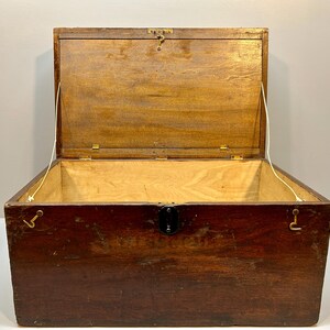 May include: An open, antique wooden chest with a hinged lid. The chest is made of dark brown wood with a lighter wood interior. It features metal hardware, including a latch and hinges. The lid is held open by a white cord.