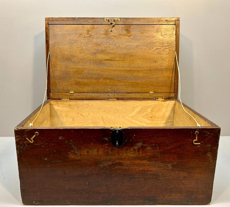 May include: An open, antique wooden chest with a hinged lid. The chest is made of dark brown wood with a lighter wood interior. It features metal hardware, including a latch and hinges. The lid is held open by a white cord.